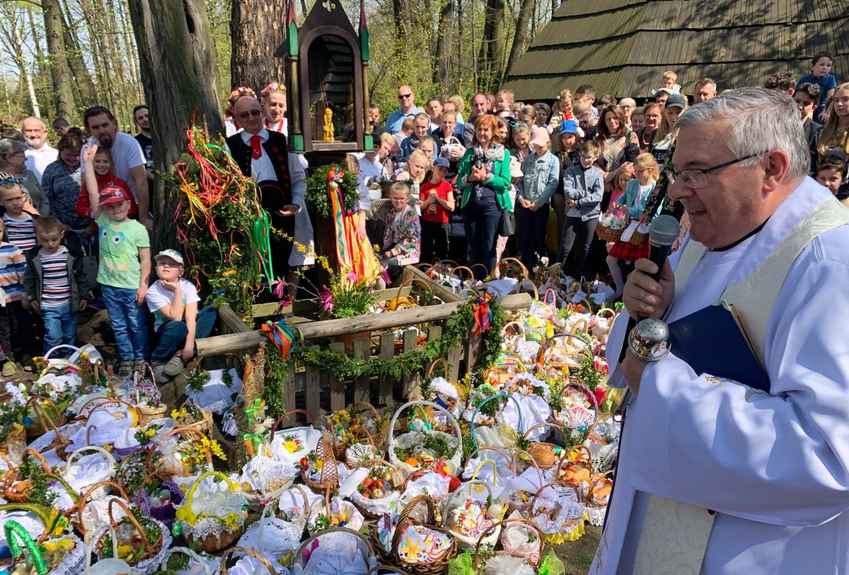 Ksiądz święcący koszyczki ułożone na ziemi. Wokół ludzie i drewniane chaty.