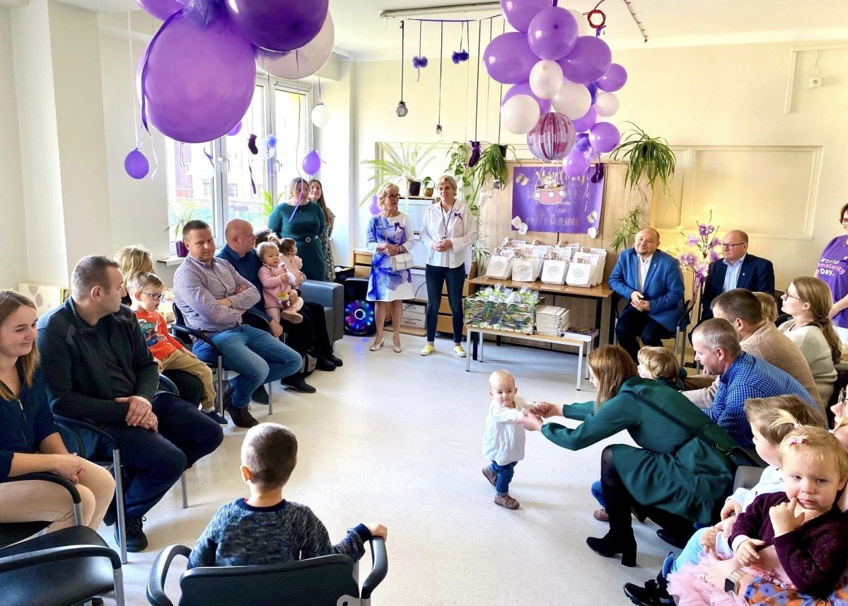 uczestnicy spotkania siedzą w okręgu, sala przyozdobiona jest na fioletowo.