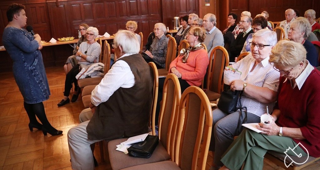 Wykład na dużej sali. Na krzesłach siedza seniorzy i słuchają prelegentki.