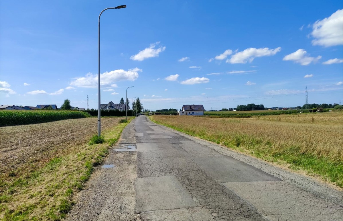 Wąska asfaltowa droga z popękaną i podziurawioną nawierzchnią, wzdłuż drogi pola.