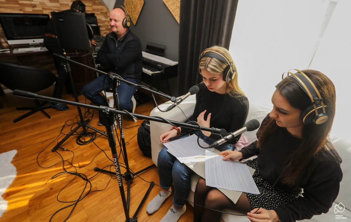 Uczennice w studiu nagrań, obok Adam Maćkowski. Nagrywają dźwięki do podcastów przy mikrofonach.