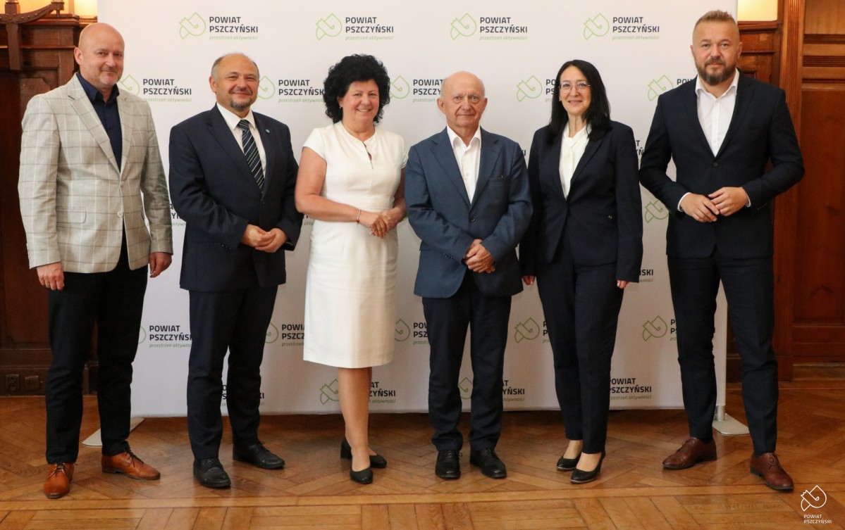 Samorządowcy pozują do wspólnego zdjęcia w sali sesyjnej starostwa na tle białej ścianki z zielonym logo powiatu pszczyńskiego.