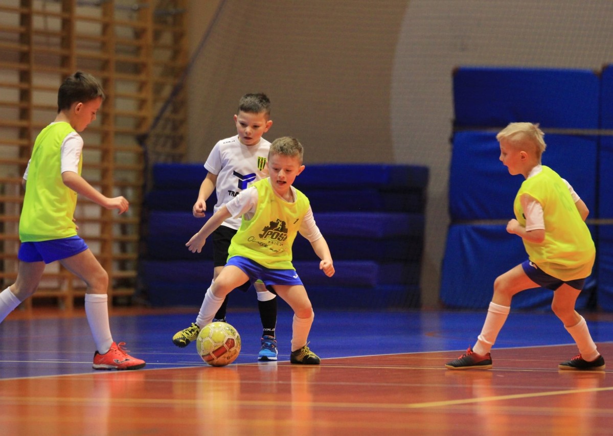 Czterech zawodników. Jeden z nich kopie do piłki. Pozostali piłkarze chcę odebrać mu piłkę. Hala sportowa. Zawodnicy mają na sobie sportowe stroje i buty.