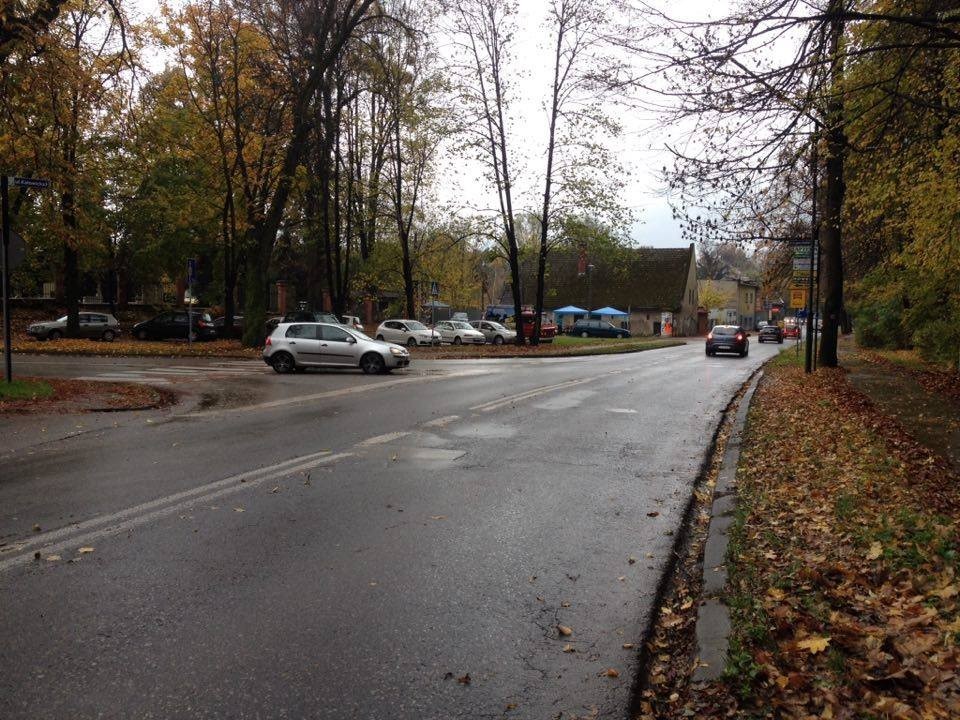 Grafika przedstawia drogę i wylot z drogi bocznej, gdzie póbuje włączyć sie do ruchu pojazd. W tle widoczne jest wejście na cmentarz.