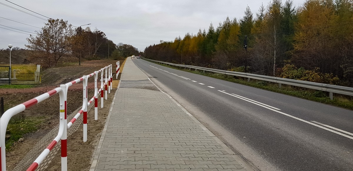 Zdjęcie przedstawia drogę, nowy fragment chodnika z kostki brukowej. Po prawej stronie znajdują się biało-czerwone barierki ochronne, a po lewej kolorowe jesienne drzewa.