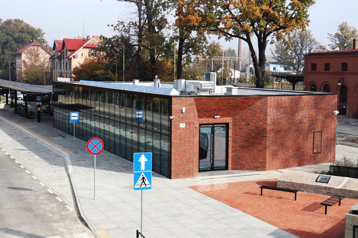 Zdjęcie przedstawia widziany z lotu ptaka budynek z pomarańczowej cegły. To pszczyńskie centrum przesiadkowe. W oddali widać wiaty autobusowe oraz budynek dworca kolejowego.