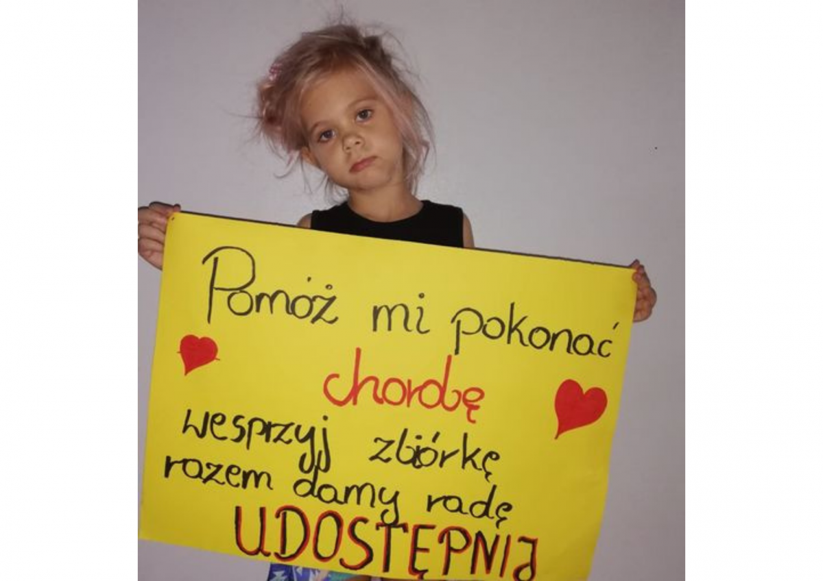 Na zdjęciu widać dziewczynkę ze smutną miną. Trzyma w dłoniach żółtą kartkę z napisem: "Pomóż mi pokonać chorobę. Wesprzyj zbiórkę. Razem damy radę. Udostępnij".
