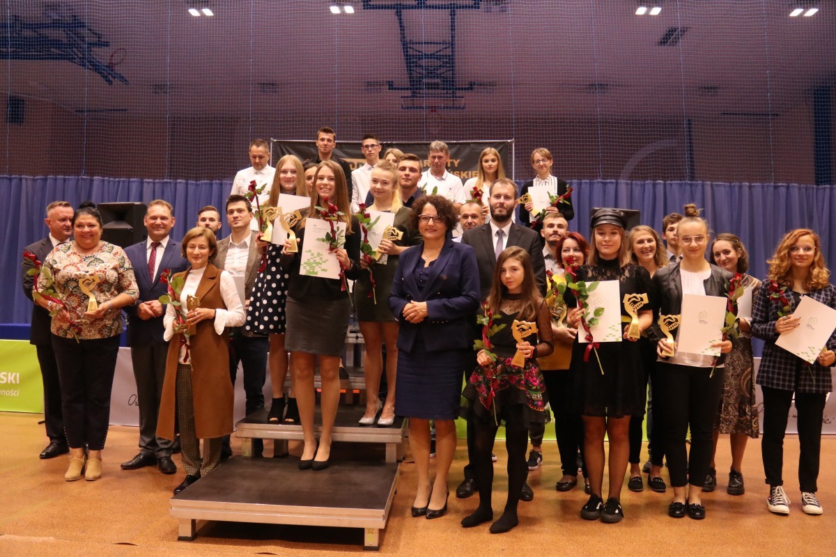 Zdjęcie grupowe przedstawia laureatów Nagrody Starosty Pszczyńskiego w dziedzinie sportu wraz z starostą, wicestarostą i dyrektorem POSiR w Pszczynie. Nagrodzeni stoją na scenie w kilu rzędach, a w tle widać siatkę i halę sportową..
