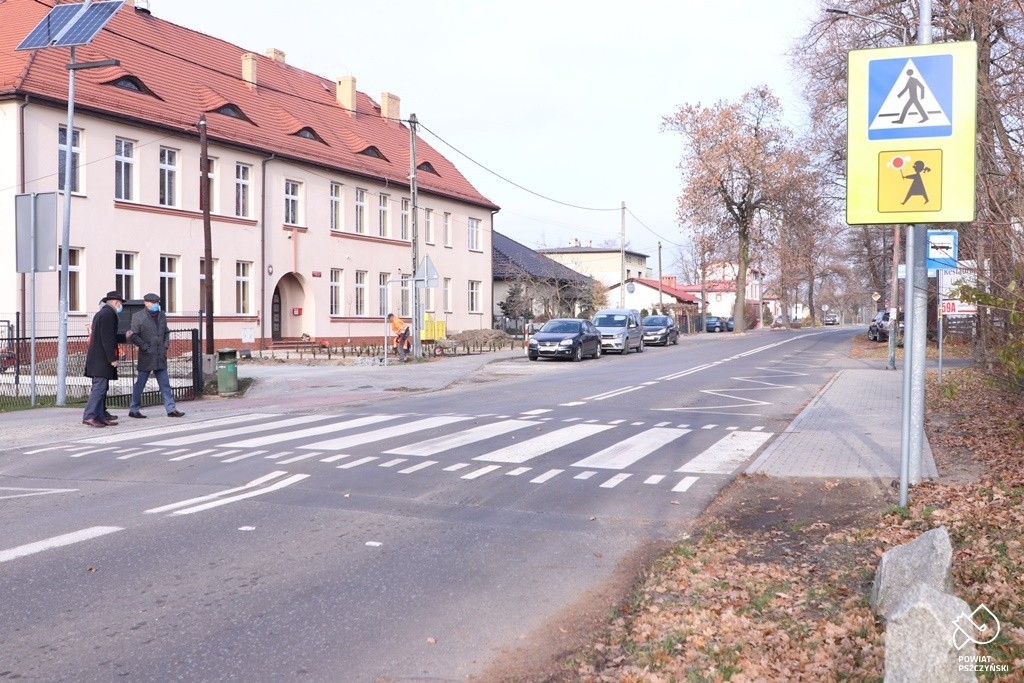 Wyniesione przejście dla pieszych przy szkole podstawowej. Po lewej stronie znajdują się znaki drogowe, u wejścia na przejście znajdują się trzy osoby.