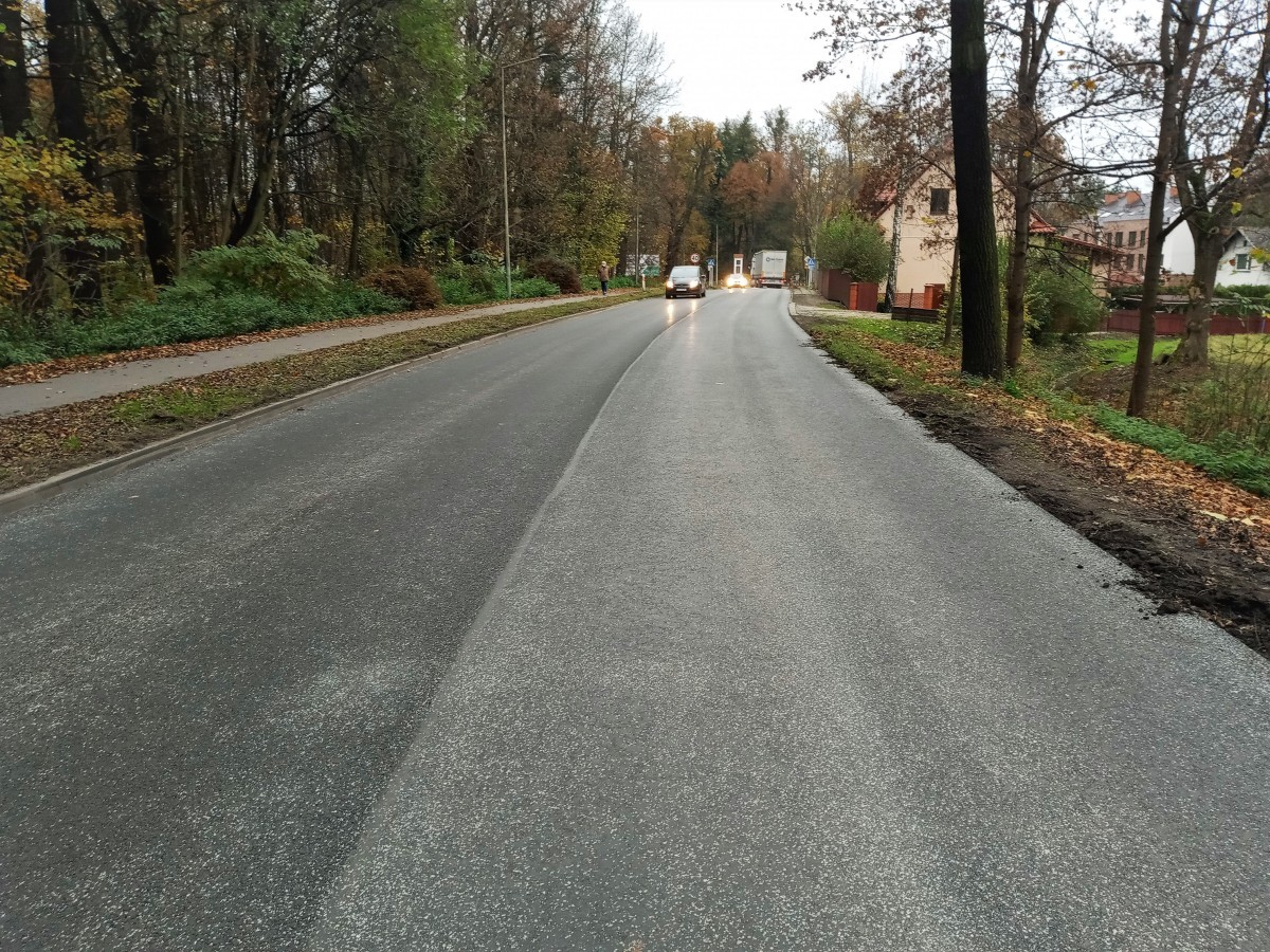 Zdjęcie przedstawia naprawioną jezdnię. Po prawej stronie znajduje się pobocze z ziemi, a po lewej chodnik dla pieszych. W tle widać kapliczkę i pszczyński park.