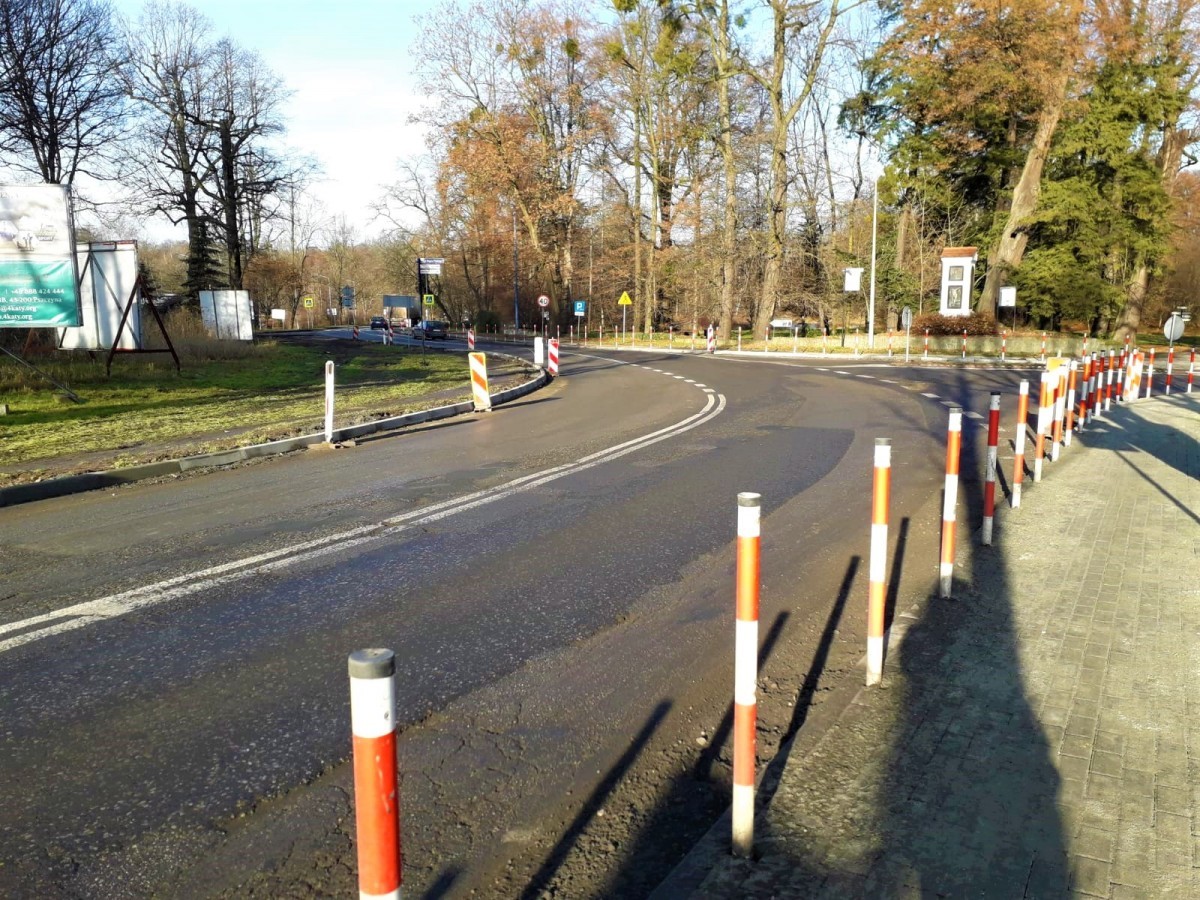 Droga wzdłuż niej chodnik i biało-czerwone słupki. W tle kapliczka i park.