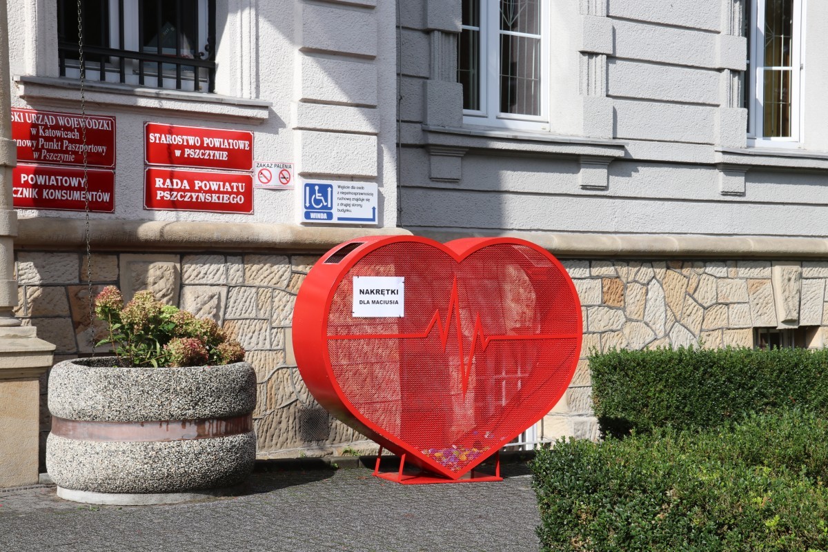 Na zdjęciu widać duże czerwone serce wykonane z siatki. Pojemnik na nakrętki stoi przed budynkiem Starostwa Powiatowego w Pszczynie.