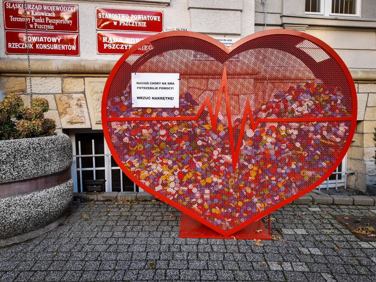 Na zdjęciu widać duże czerwone serce wykonane z siatki. Pojemnik na nakrętki stoi przed budynkiem Starostwa Powiatowego w Pszczynie. Serce jest do połowy wypełnione kolorowymi, plastikowymi nakrętkami.