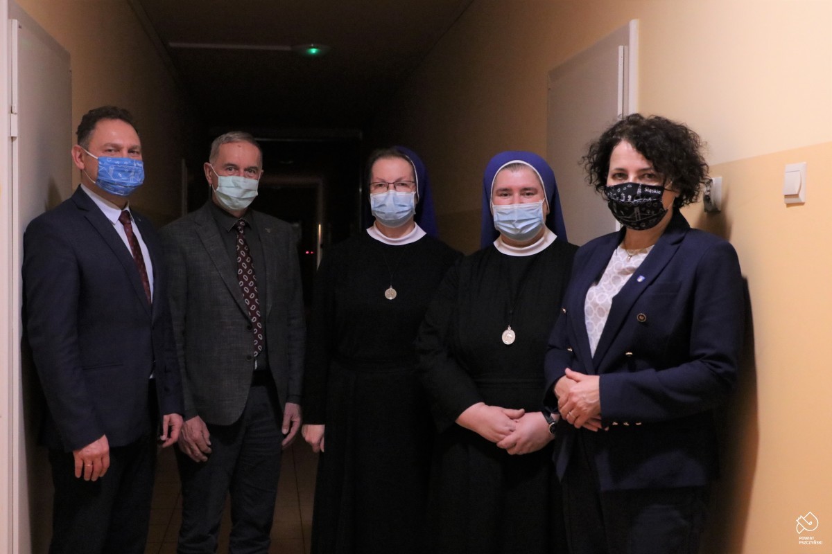 Zdjęcie przedstawia dwie siostry zakonne oraz troje przedstawicieli Zarządu Powiatu Pszczyńskiego. Stoją obok siebie w ciemnym korytarzu. Na twarzach mają założone maseczki.