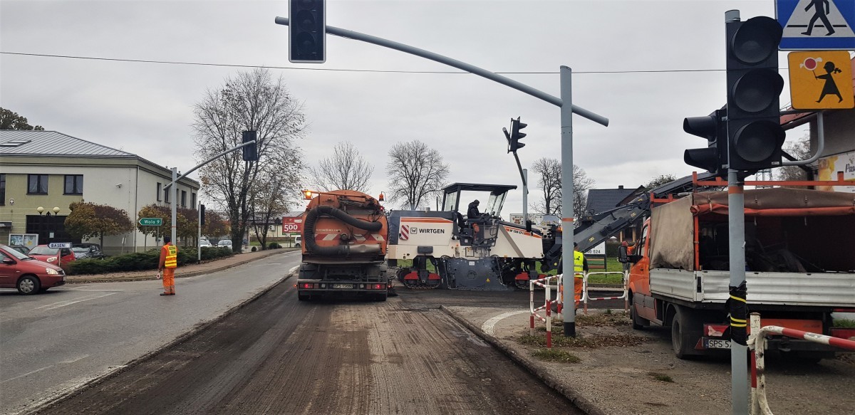 Zdjęcie przedstawia skrzyżowanie, na którym pracują maszyny. Z jednej części jezdni ściągnięty jest asfalt. Widać także pracowników kierujących ruchem oraz samochody.