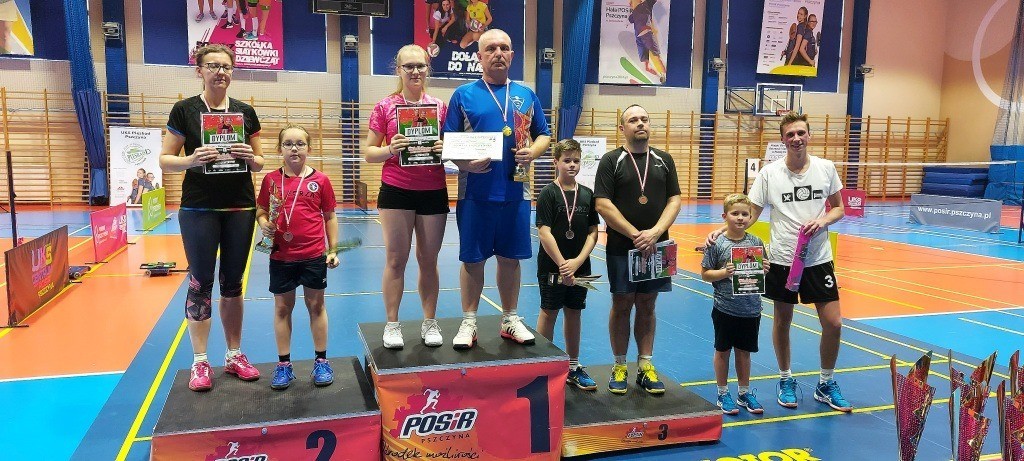 Na podium w hali sportowej stoją uczestnicy turnieju. W dłoniach trzymają dyplomy i puchary, a na szyjach mają zawieszone medale. Ubrani są w sportowe stroje. W tle widać drewniane drabinki.