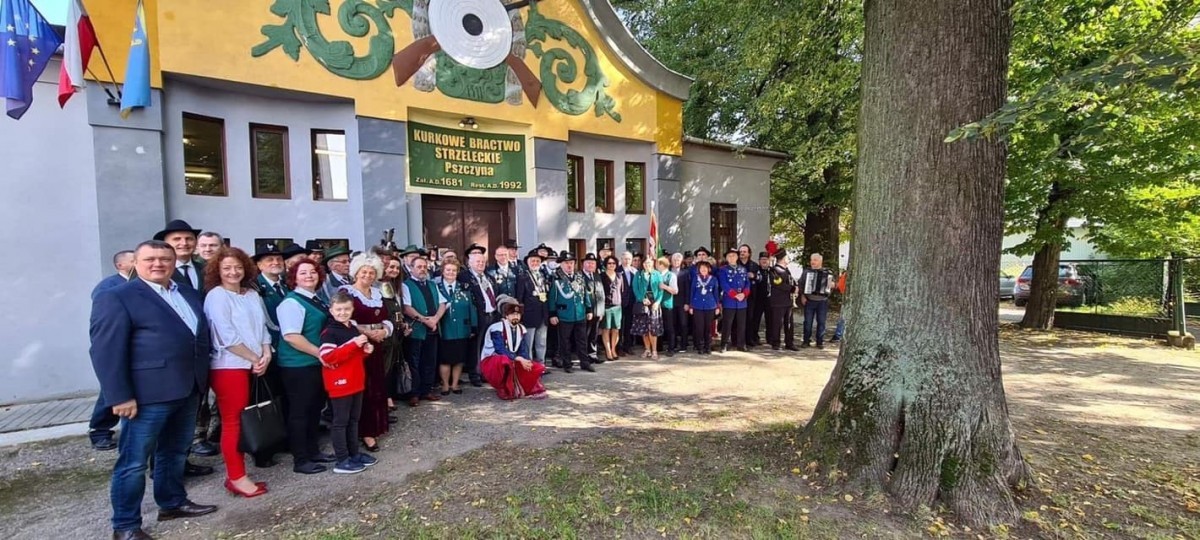 Zdjęcie przedstawiające uczestników zawodów strzeleckich stojących w kilku rzędach przed szarym budynkiem strzelnicy Kurkowego Bractwa Strzeleckiego w Pszczynie. Na pierwszym planie widać pień drzewa, a w tle korony drzew.