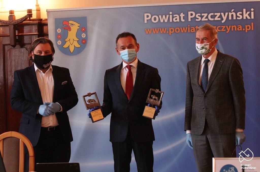 Grafika przedatawia trzech mężczyzn, ubranych w maseczki i rękawiczki mężczyzna w środku trzyma dwie statuetki żubra pszczyńskiego