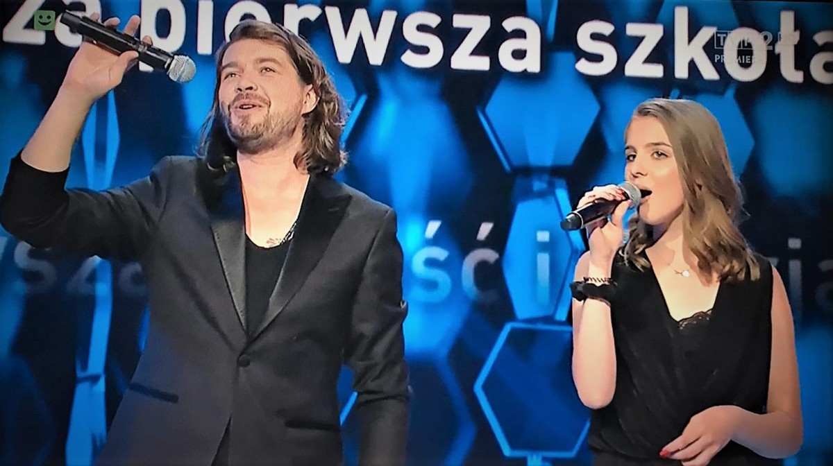 Zdjęcie przedstawia dwóch wokalistów. Jednym z nich jest gwiazda grupy Pectus oraz dziewczyna, która wygrała odcinek programu telewizyjnego Szansa na sukces. W tle widać niebieskie światła i słowa tekstu piosenki, którą wykonali wspólnie artyści.