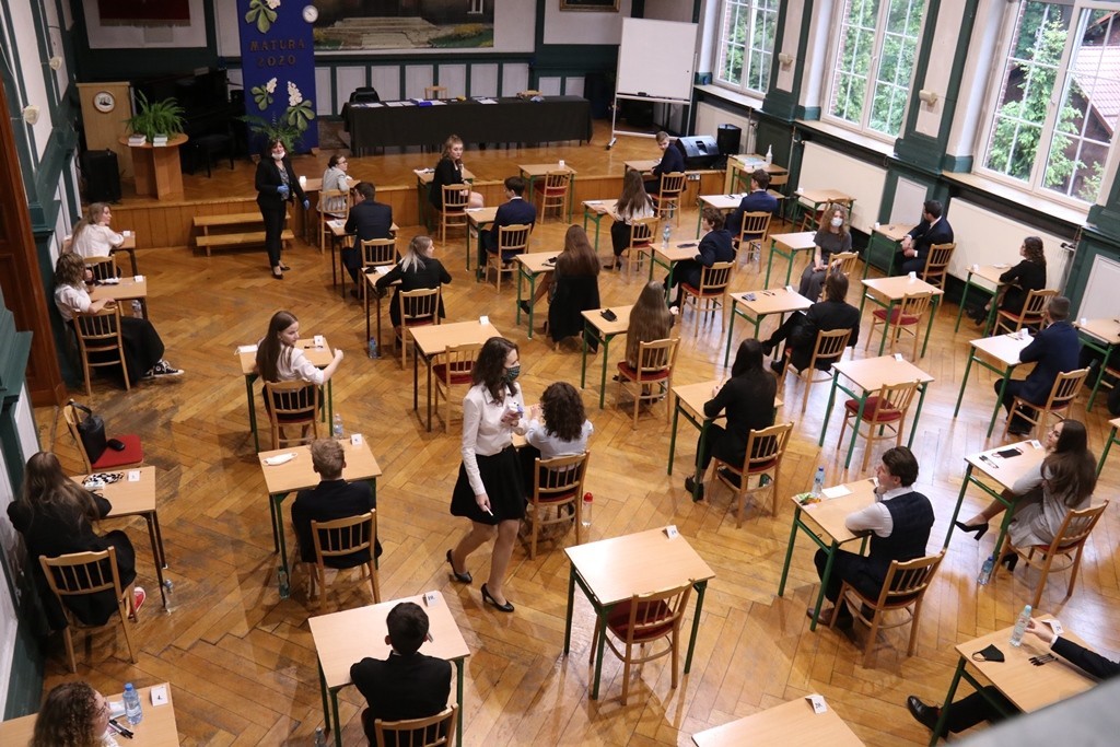 Aula szkolna. W rzędach ustawione ławki i krzesła, na których siedzą maturzyści przed egzaminem maturalnym.