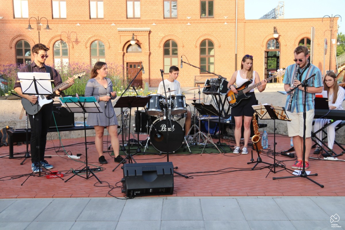 Młodzieżowy zespół Offbeat gra koncert na placu przy centrum przesiadkowym w Pszczynie. W tle budynek dworca kolejowego.