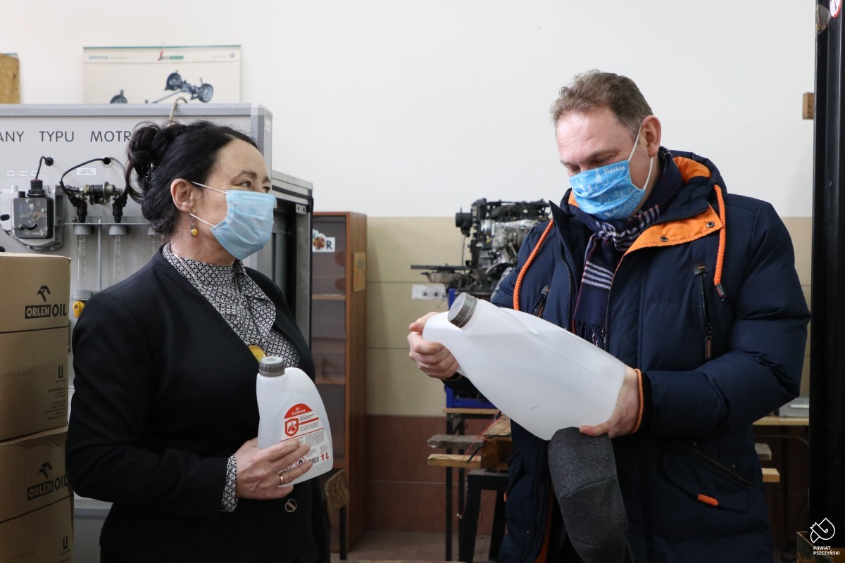 Kobieta i męzczyzny znajduja się w pracowni samochodowej, trzymają w rękach duże pojemniki z płynem do dezynfekcji.