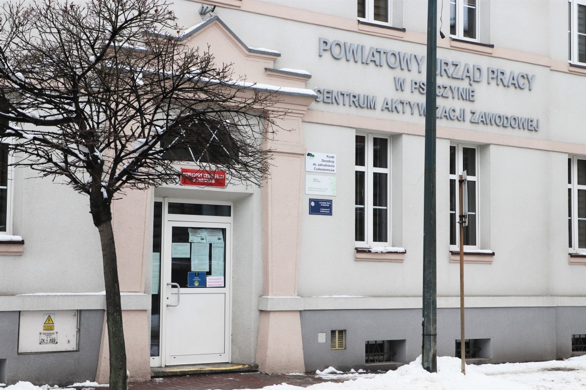 Szary budynek z duża ilością okien. Na budynku napis: Powiatowy Urząd Pracy w Pszczynie. Przed budynkiem odśnieżony chodnik i drzewa. Zima.