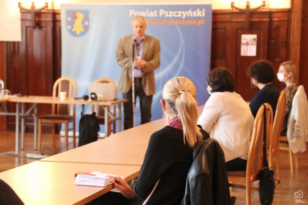 Duża sala, za stołami ułożonymi w kwadrat siedzą cztery kobiety, na dalszym planie stoi mężczyzna z białą laską. Prowadzi szkolenie
