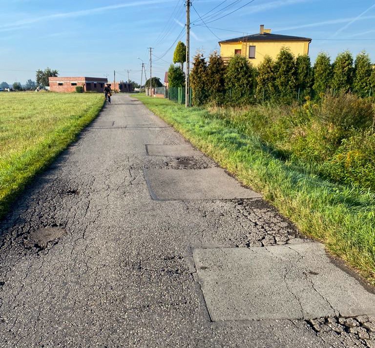 Droga z dziurami, obok pola i domy.