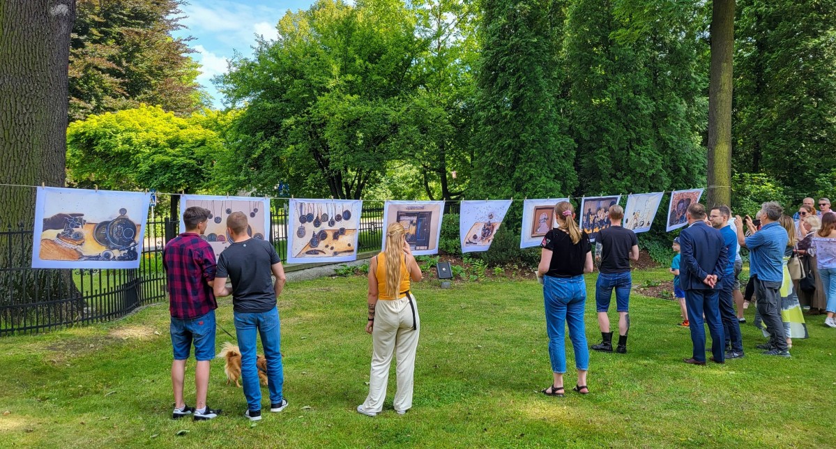 Grupa ludzi stoi tyłem w ogrodzie starostwa i ogląda obrazy wywieszone na sznurze jak pranie