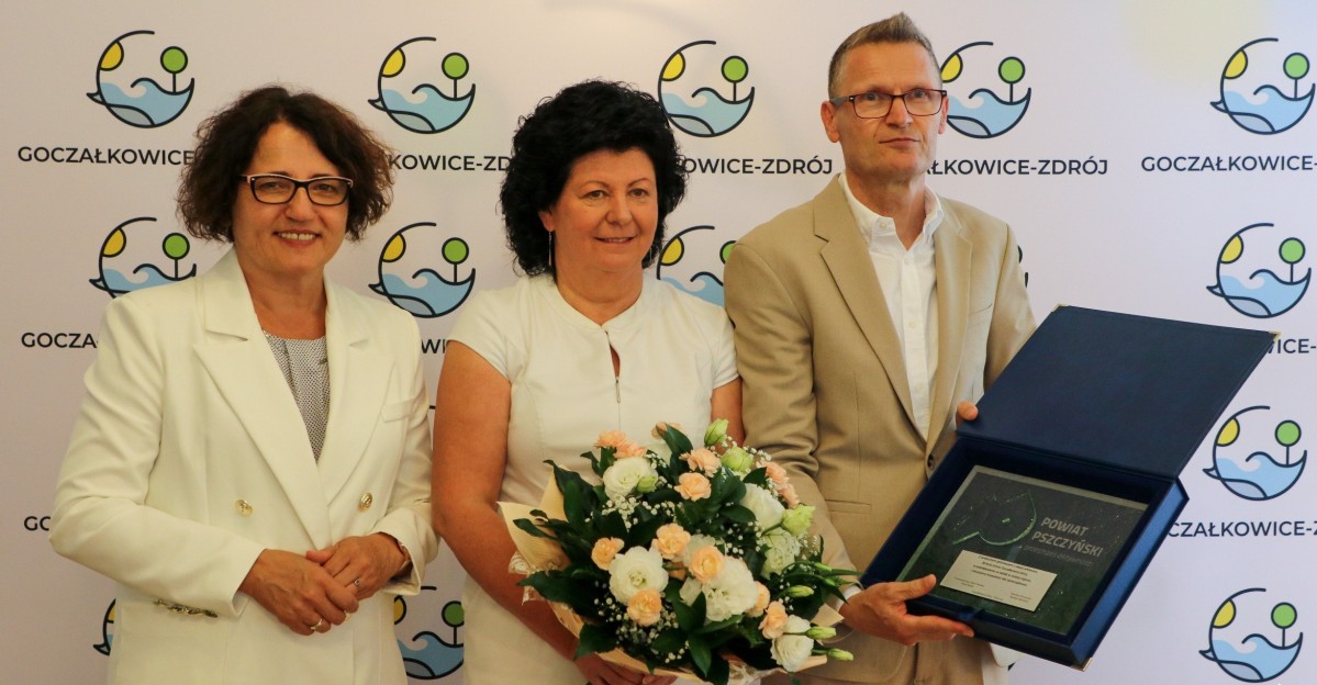 Starosta, wójt gminy Goczałkowice-Zdrój z kwiatami i przewodniczący Rady Gminy trzymający grawer na tle ścianki gminnej.